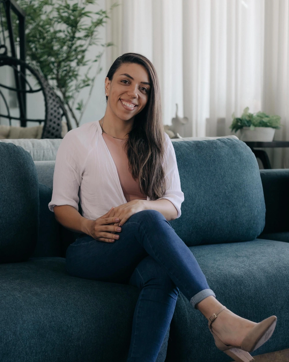 Bruna Chagas sorrindo, sentada de pernas cruzadas em um sofá azul, de camisa clara e calça jeans